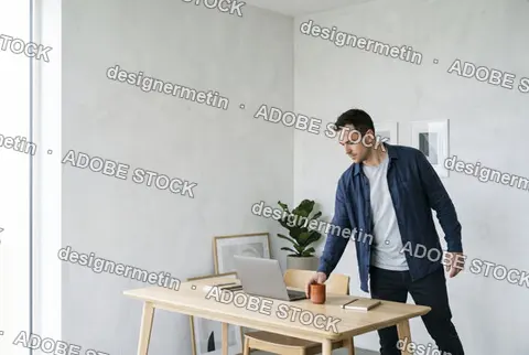 Young Man Working Remotely On A Laptop Standing By A Modern Wooden Desk In A Bright Minimalist Room